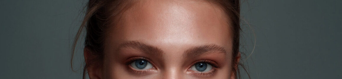 Nærbillede af en kvinde med naturligt makeuplook: blå øjne, jævn hud med varm bronzeblush, diskret øjenskygge, definerede bryn og blanke nude læber. Håret er samlet i en løs knold med enkelte løse totter. Billedet har et afdæmpet gråt baggrund og blødt, ensartet lys, der fremhæver ansigtets konturer og hudens glød. Perfekt til skønheds- eller kosmetikkommunikation.