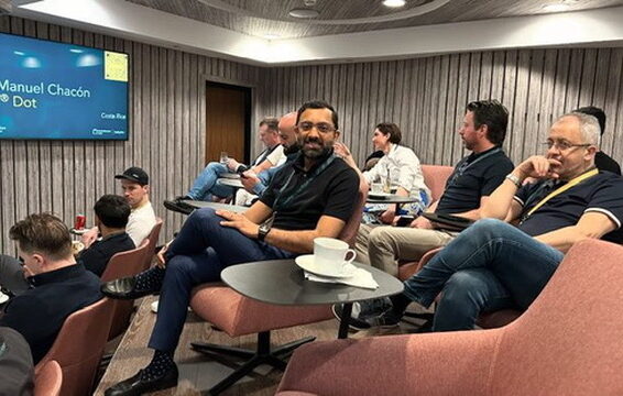 Et mødelokale med flere personer siddende i rosa polstrede loungesæder omkring små, sorte runde sideborde. På et bord står en hvid porcelænskop på en underkop, og flere deltagere bruger bærbare computere, notesbøger og mobiltelefoner. Et stort vægmonteret display viser en blå præsentationsskærm. Rummet har træpaneler og dæmpet belysning, der skaber en afslappet konferenceatmosfære.