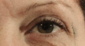Nærbillede af et enkelt øje med en grønbrun iris, tydelige øjenvipper og let øjenmakeup langs den øverste vippekant. Et velplejet øjenbryn med naturlig form ses øverst, og hudens struktur med fine linjer og lette skygger er tydelig. Der er en diskret skygge ved ydre øjenkrog. Billedet fokuserer på øjets detaljer, kontraster og nuancer i hud- og øjenfarve i et varmt, naturligt lys.