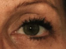 Nærbillede af et enkelt øje i grønne nuancer, med tydelig refleks i iris og sort pupil. Øjet er markeret med mørk øjenvippebørstning og mascara, samt en let sort eyeliner langs øjenkanten. Huden omkring viser naturlig tekstur, fine linjer og en brunlig hudtone, mens delen af et bryn og øvre øjenlåg skimtes i billedets tætte beskæring.
