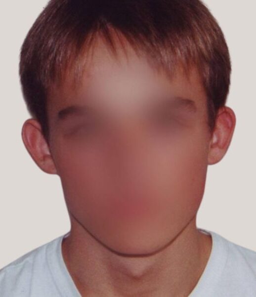 Portræt af en person med sløret ansigt og kort brunt hår mod en neutral, lys baggrund. Fokus på tøjet: en enkel hvid t‑shirt med rund hals og glat, uprintet stof, der giver et afslappet og neutralt udtryk. Billedet er tæt beskåret om hoved og skuldre, hvilket fremhæver t‑shirtens rene linjer og dagligdags, ungdommelige stil.