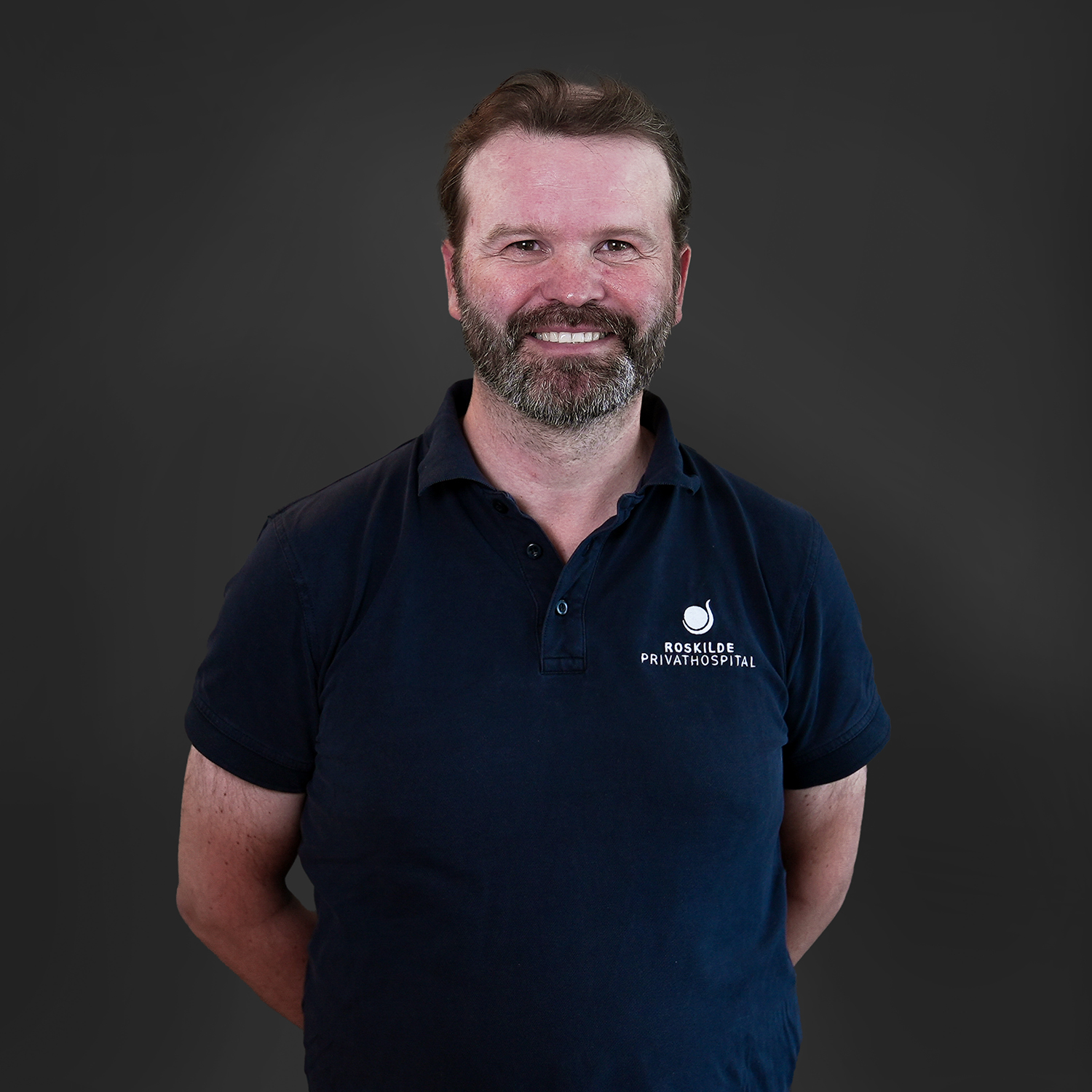 En mand iført en mørkeblå poloshirt med korte ærmer og knappet krave står mod en neutral grå baggrund. På venstre bryst ses et hvidt logo og teksten "Roskilde Privathospital", tydeligt synlig. Skjorten har en afslappet pasform og et rent, professionelt udtryk, der fremhæves af den enkle farvekontrast mellem det mørke stof og det lyse logo.