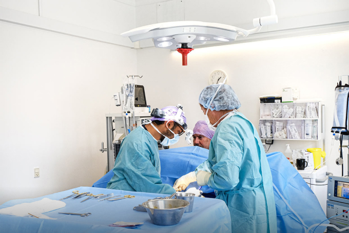 Operativt billede fra operationsstue på Roskilde Privathospital: to kirurger i sterile kit arbejder koncentreret ved patientens bryst under blå kirurgiske dække, kirurgisk lyskilde over feltet, instrumenter og bakker på bord, sterile handsker og roligt arbejdsmiljø — brystforstørrende operation (BFO) udført med professionel omhu og 30 års erfaring.